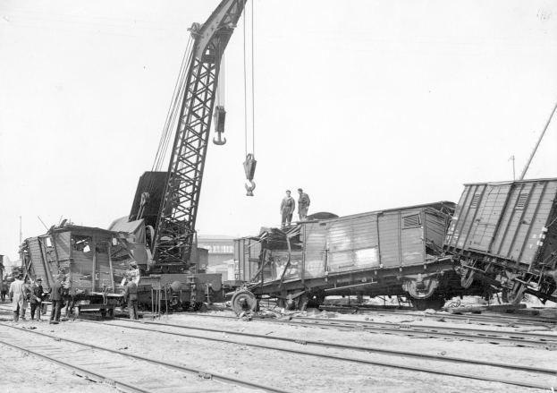 Schwarzes und weißes Bild von einer Zugkatastrophe mit einem Kran, der ein Waggon auf die Schienen hebt, mehrere Menschen in der Nähe und im Hintergrund Gebäude und einen klaren Himmel.