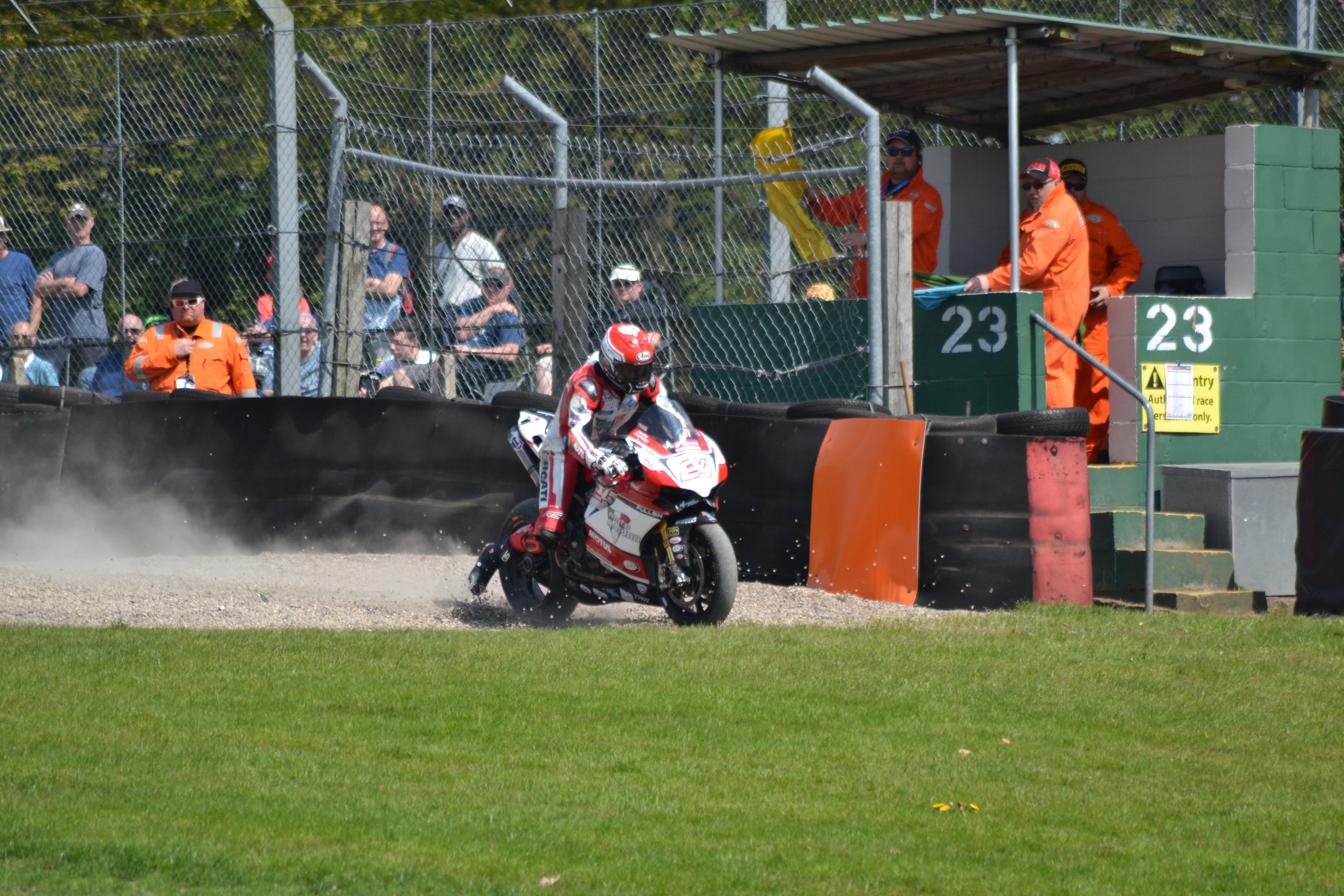 Ein Motorradfahrer mit Helm und Handschuhen fährt über ein grasbewachsenes Feld, mit Zuschauern in Mützen und Schutzbrillen, einem eingezäunten Bereich, Treppen mit Geländern und einem nummerierten Schuppen im Hintergrund.