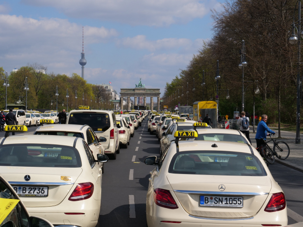 Eine lange Reihe von Taxis, die an einer belebten Straße in Berlin, Deutschland, geparkt sind, mit Fahrradfahrern und Fußgängern auf dem Gehweg, flankiert von Bäumen und Laternenmasten und Gebäuden, einem Bogen und einem Turm im Hintergrund bei einem bewölkten Himmel.
