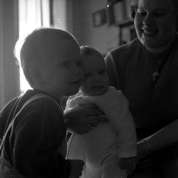 Eine Frau lächelt warm, während sie ein Baby hält, mit zwei anderen Kindern neben ihr in einem gemütlichen Raum mit Rahmen und einem Fenster im Hintergrund.