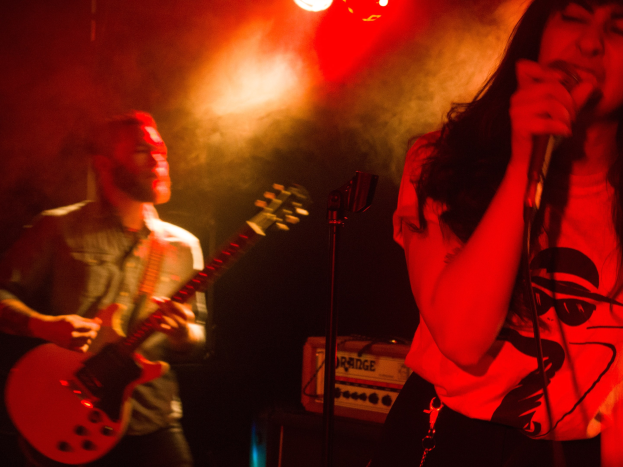 Eine Frau singt in ein Mikrofon auf der Bühne, während ein Mann im Hintergrund eine akustische Gitarre spielt und die Deckenbeleuchtung die Szene erhellt.