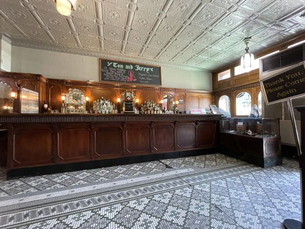 Ein großer Raum mit einer zentralen Bar, Deckenlampen, Flaschen auf einem Tisch und einer Tafel mit Text auf der rechten Seite, befindet sich im Tom and Jerry Pub in London.