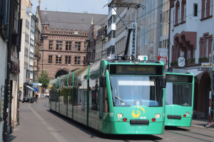 Zwei grüne Straßenbahnen fahren auf einer von hohen Gebäuden gesäumten Stadtstraße, mit parkenden Fahrrädern und Fußgängern auf den Gehwegen, unter einem klaren blauen Himmel mit einem Baum im Hintergrund.