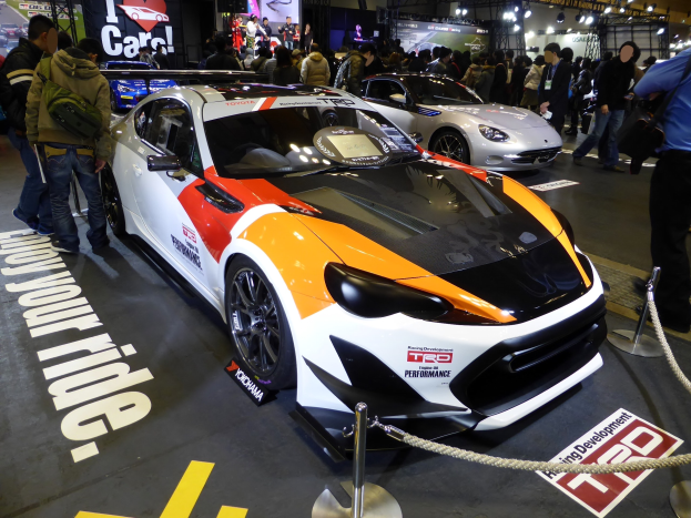 Eine Gruppe von Menschen steht um ein Rennfahrzeug herum, das auf der Tokyo Motor Show ausgestellt ist, umgeben von Absperrpollern mit Seilen, mit Text- und Bildtafeln im Hintergrund unter beleuchteter Decke.