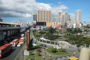 Eine belebte Stadtstraße mit verschiedenen Fahrzeugen, Gebäuden, einer Brücke und Grün unter einem bewölkten Himmel.