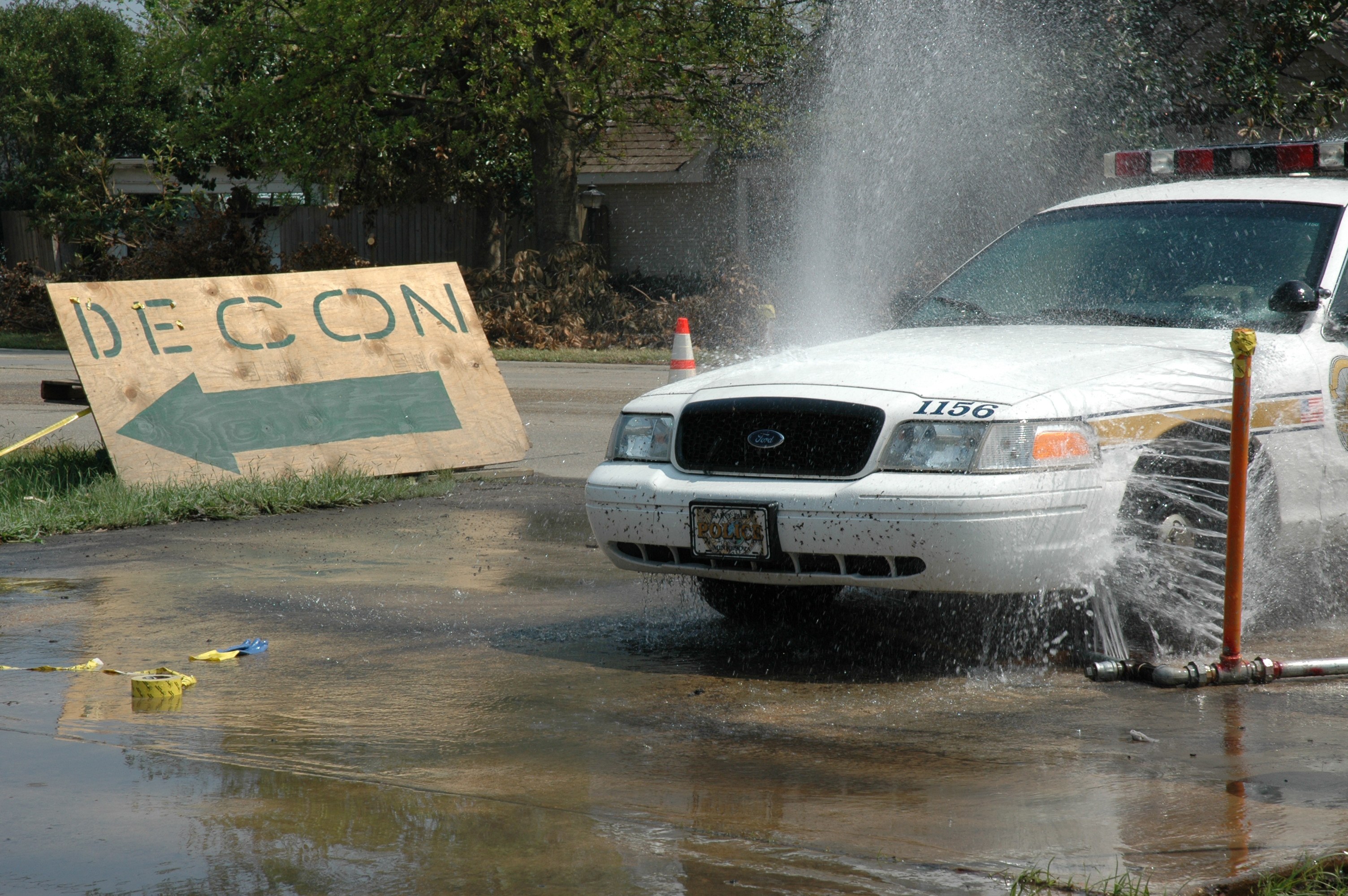 Ein Streifenwagen fährt durch eine Pfütze, mit einem Schild, einer Verkehrskegel und anderen Gegenständen auf dem Boden, mit Bäumen, Gebäuden und einem klaren blauen Himmel im Hintergrund.