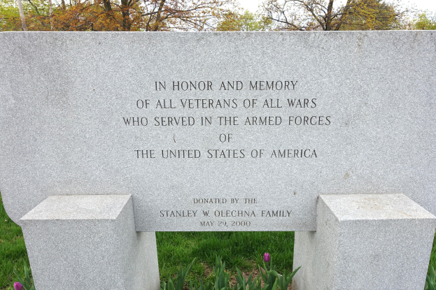 Ein Gedenkstein mit der Inschrift "In Ehren und Andenken aller Kriegsveteranen aller Kriege, die in den Streitkräften der Vereinigten Staaten von Amerika dienten" umgeben von Pflanzen, Blumen, Gras, Bäumen und einem bewölkten Himmel.