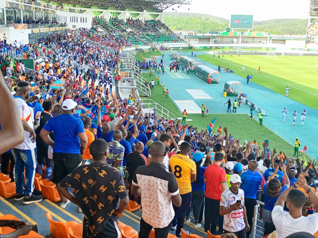 Eine große Menschenmenge versammelt sich vor einem eingezäunten Fußballfeld mit Hügeln und einem klaren blauen Himmel im Hintergrund und erwartet das Spiel der afrikanischen Nationenmeisterschaft 2019.
