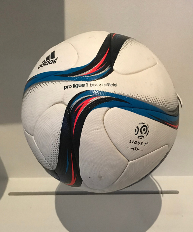Ein weißer Fußball mit dem Adidas-Logo und der Aufschrift 'Ligue 1' liegt auf einem weißen Tisch gegen einen hellen weißen Hintergrund.