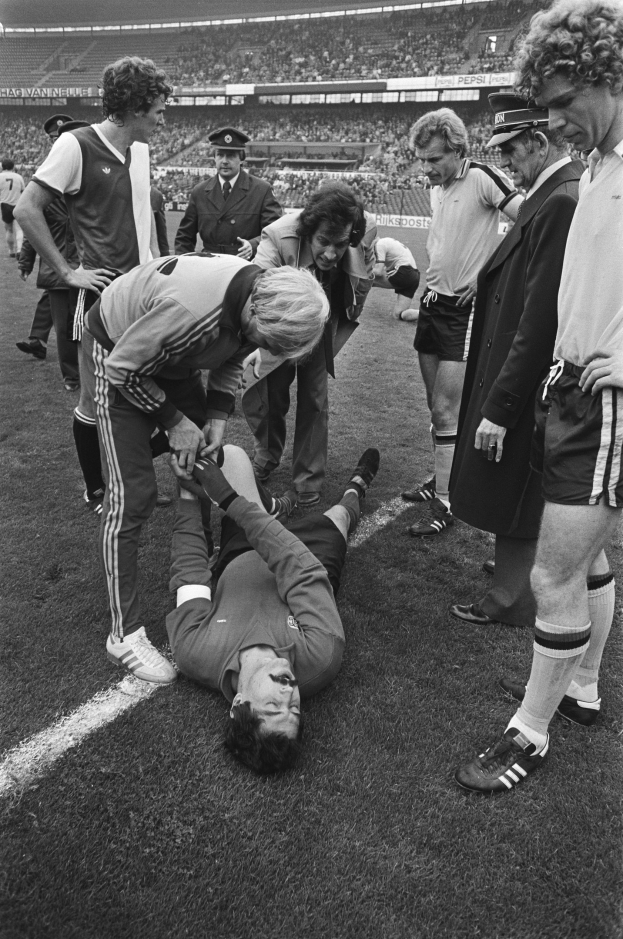 Schwarzes Bild eines Fußballspielers, der auf dem Boden liegt, während ihn eine Gruppe von Männern und eine Stadionmenge umgibt, was darauf hindeutet, dass er verletzt sein könnte.