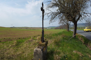 Ein gelbes Auto fährt auf einer Straße neben einem Baum entlang, mit einem Kreuz, das die Stelle eines tödlichen Autounfalls markiert, vor einem Hintergrund aus Hügeln, Wolken und grüner Wiese mit Blumen.
