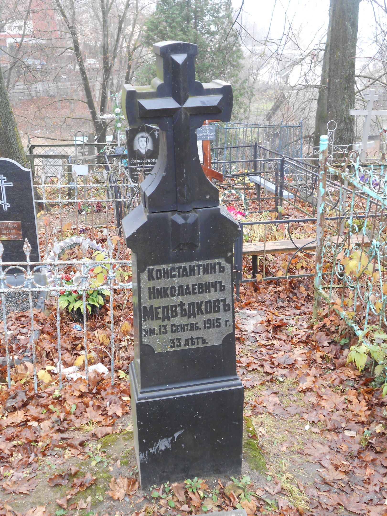 Ein Friedhof mit einem zentralen Kreuz, umgeben von Grabsteinen, Eisenzäunen, Pflanzen, Bäumen und fernen Gebäuden, bedeckt mit trockenen Blättern.