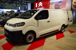 Ein weißer Citroën Berlingo Van wird auf einer Autauschau präsentiert, umgeben von schwarzer Schlafzimmer-Möblierung, während die Wände mit Brettern und Lichtern verziert sind.