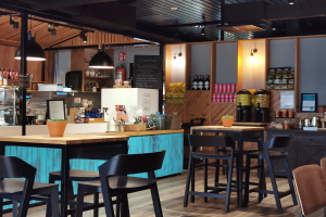 Innenraum eines Restaurants mit Holztischen, Stühlen, einer Topfpflanze auf einem Tisch, einer Schiefertafel, einem Kühlschrank, einem Feuerlöscher, Regalen mit Flaschen, Schränken mit Gegenständen, einem Rahmenfoto an der Wand und Deckenleuchten.