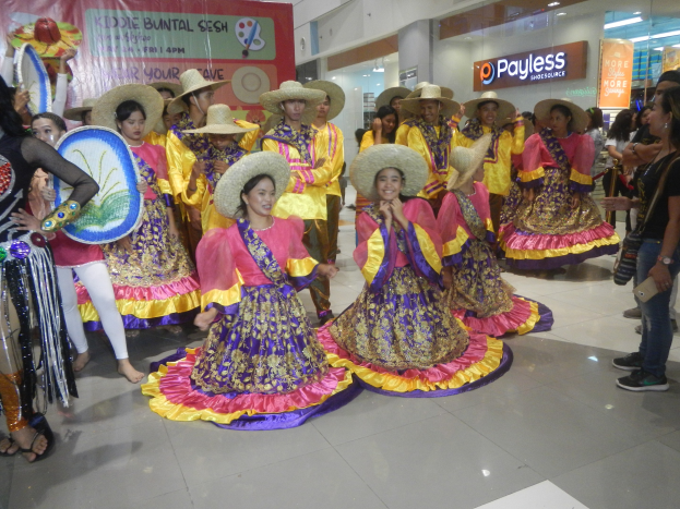 Eine Gruppe von Menschen in bunten Kleidern und Hüten tanzt in einem Einkaufszentrum, einige stehen und halten Musikinstrumente, ein Banner mit Text und festliche Deko wie Deckenlichter und Schilder mit Text.