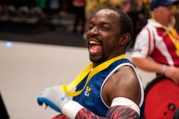 Ein Mann im Rollstuhl mit ausgestreckten Armen und einer Medaille um den Hals lächelt im Vordergrund, während im Hintergrund Menschen, Stühle und andere Gegenstände zu sehen sind.
