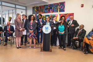 Gruppe von Menschen in einem Raum mit einem Podium, einem Mikrofon und einer Tafel mit dem Text "Sancho-Stadtrat tritt dem Stadtrat bei", umgeben von Fahnen, Stühlen und einem Feuerlöscher an der Rückwand.