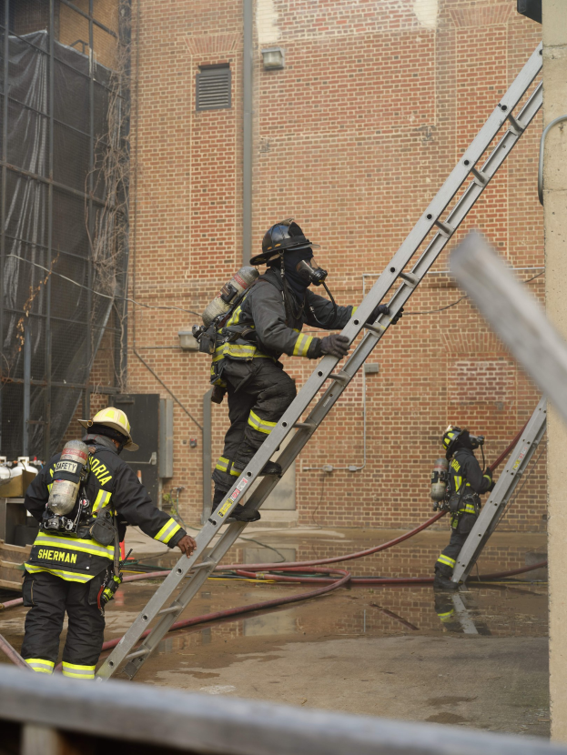 Feuerwehrleute mit Helmen und Luftzylindern klettern an einer Leiter vor einem Backsteingebäude hoch, mit Rohren auf dem Boden und einer Metallstange am Boden; im Hintergrund ist ein Gebäude mit Fenstern und Netz zu sehen.