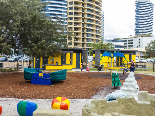 Spielplatz in städtischer Umgebung mit Kindern auf Geräten, umgeben von Bäumen und hohen Gebäuden, Fahrzeugen auf der Straße und sichtbarem Himmel.