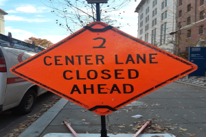 Orange "2 Center Lane Closed Ahead" Schild an der Stra├če mit Fahrzeugen, Masten, Planken, B├Ąumen, Geb├Ąuden und einem bew├Âlktem Himmel im Hintergrund.