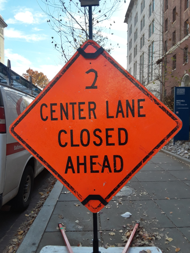 Orange "2 Center Lane Closed Ahead" Schild an der Stra├če mit Fahrzeugen, Masten, Planken, B├Ąumen, Geb├Ąuden und einem bew├Âlktem Himmel im Hintergrund.