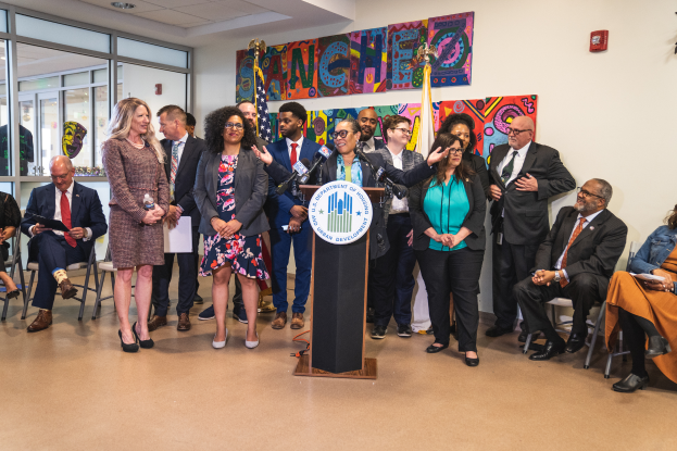Eine Gruppe von Menschen steht und sitzt in einem Raum mit einem Podium, einem Mikrofon und einer Tafel mit der Aufschrift "Sancho-Stadtrat tritt dem Stadtrat bei", mit Flaggen, einer Tafel, einem Feuerlöscher und Deckenleuchten an der Rückwand.