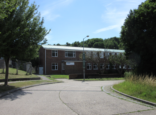 Ein Schulgebäude mit Fenstern, ein Straßenschild, ein Schild, eine Baumgruppe, Gras, ein Zaun und ein bewölkter Himmel, umgeben von saftigem Gras.