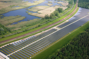 Luftaufnahme einer Solaranlage mit Panelen, umgeben von Bäumen, Gras, Wasser und einer Bahn auf einem Gleis.