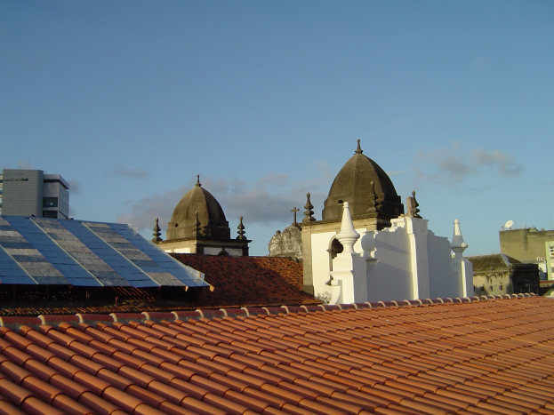 Eine Stadtansicht mit mehreren Gebäuden im Vordergrund unter einem blauen Himmel, mit Solarpanelen auf einem Dach.