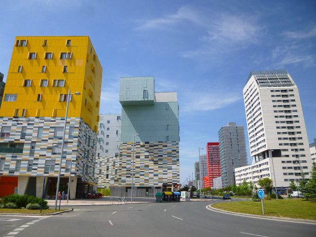 Eine Stadtstraße mit hohen Gebäuden im Hintergrund, Straßenlaternen, Autos, Infotafeln, Büschen, Bäumen und einer bewölkten Himmel, mit einzigartig gestalteten Gebäuden der Universität Rotterdam.