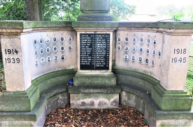 Ein Holocaust-Denkmal in einem jüdischen Friedhof in Berlin, mit einer Tafel mit Text und Zahlen an seiner Wand, umgeben von Bäumen, einem Zaun und trockenen Blättern auf dem Boden.