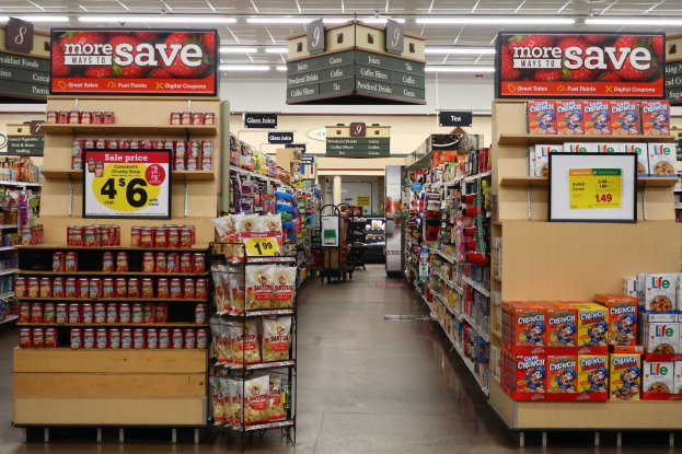 Ein Lebensmittelgeschäft mit Regalen und Boden voller verschiedener Lebensmittel, beleuchtet von Deckenlampen und mit Informationsschildern im Hintergrund und einer Person im Hintergrund.