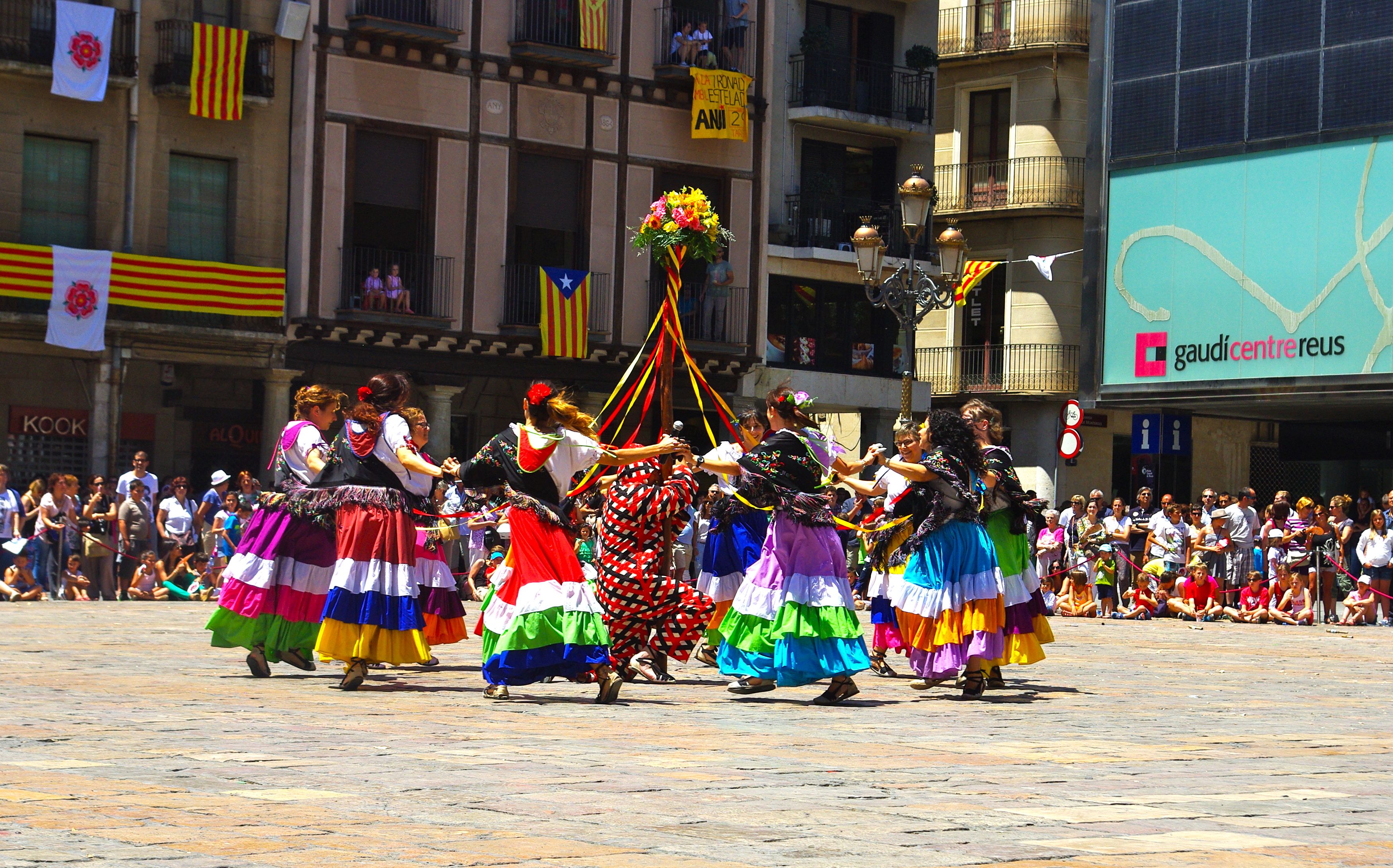 Eine Gruppe von Frauen in bunten Köthern tanzt auf der Straße, umgeben von einer Menge Menschen, mit Gebäuden, Geländern, Balkonen, Flaggen, einem Lichtmast und einer Tafel mit Schrift im Hintergrund.