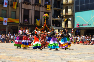 Eine Gruppe von Frauen in bunten Köthern tanzt auf der Straße, umgeben von einer Menge Menschen, mit Gebäuden, Geländern, Balkonen, Flaggen, einem Lichtmast und einer Tafel mit Schrift im Hintergrund.