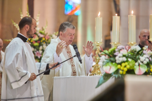 Der Bischof der Kirche des Heiligen Grabes steht an einem Rednerpult mit einem Mikrofon, spricht zu einer Gruppe von Menschen, mit einem Tisch mit Kerzen und Blumen zu seiner Rechten und Blumensträußen und einem Bleiglasfenster im Hintergrund.