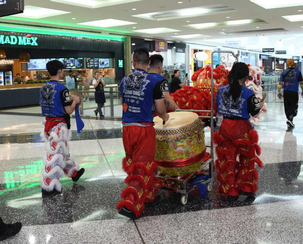 Eine Gruppe von Menschen in roter und blauer Kleidung mit Musikinstrumenten und Gepäckwagen geht durch einen hell erleuchteten Flughafen während einer Chinesischen Neujahrsfeier in Singapur.