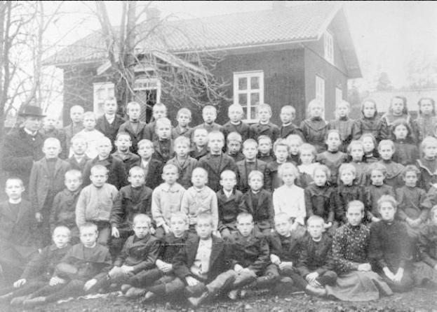 Schwarz-weiß-Foto einer Gruppe von Kindern, die vor einem Haus mit Fenstern posieren, umgeben von Bäumen und einem klaren Himmel, mit Text unten, der "1930er-Schulkinder" lautet.