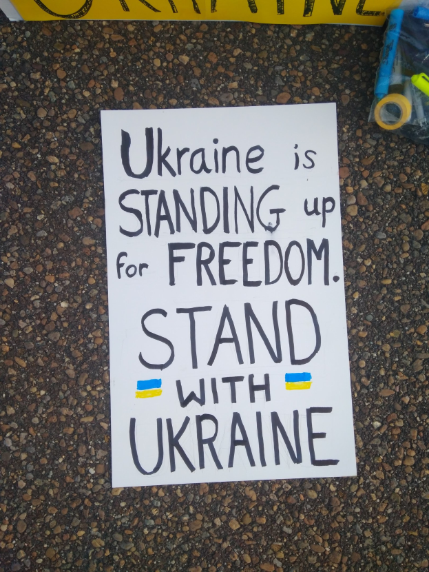 Ein weißes Papier mit der Aufschrift "Ukraine steht für Freiheit ein, steht mit Ukraine" vor einem gelben Banner, rechts daneben eine Plastikhülle mit verschiedenen Gegenständen.