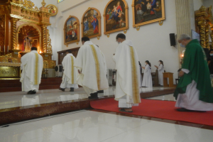 Eine Gruppe von Priestern geht eine rote Teppich in einer Kirche hinunter, mit Menschen, die auf dem Boden vor und rechts von ihnen stehen, und Fotorahmen an der Hintergrundwand.