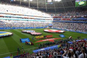 Eine große Menschenmenge in einem Stadion, die ein Fußballspiel schaut, wobei einige Fahnen und Banner halten, ein Bildschirm auf der rechten Seite und ein Mann in der Mitte umgeben von der Menge.