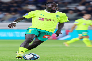 Ein Fußballer in einem gelben T-Shirt und grünen Shorts ist im Einsatz auf dem Feld, mit einer blauen Tafel und unscharfen Menschen im Hintergrund.