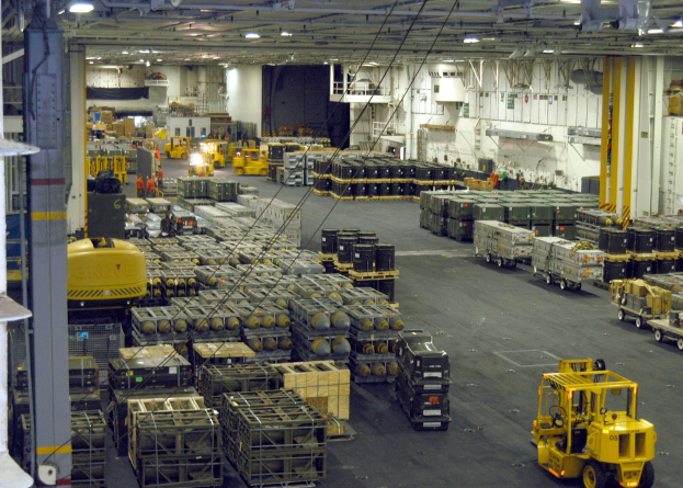 Lagerinneres mit Lkw, Containern, Menschen, Deckenleuchten, Metallstangen, diversen Gegenständen und einem sichtbaren Rad.