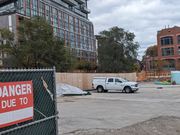 Lkw vor einem Gebäude mit einer 'Gefahr wegen offener Baustelle'-Tafel geparkt, Bäume und Gebäude im Hintergrund unter einem klaren blauen Himmel.