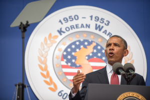 Präsident Obama, gekleidet mit Blazer, Krawatte und Hemd, hält eine Rede am Korean War Memorial in Washington, D.C., stehend hinter einem Podium mit Mikrofonen, mit einem Banner mit Text und einem Emblem dahinter und einem Ständer links von ihm.