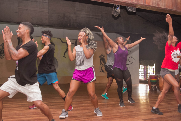 Gruppe von Menschen, die in einem Studio mit einer bemalten Wand, einem Fenster und Deckenbeleuchtung tanzen, was auf einen Zumba-Kurs hinweist.