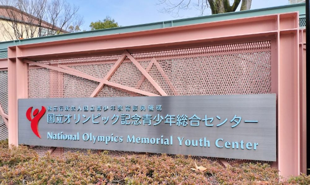 Außenansicht des Nationalen Olympischen Memorial Youth Center mit einer Wandtafel, Pflanzen am Boden, Bäumen und Gebäuden im Hintergrund und einem klaren blauen Himmel darüber.