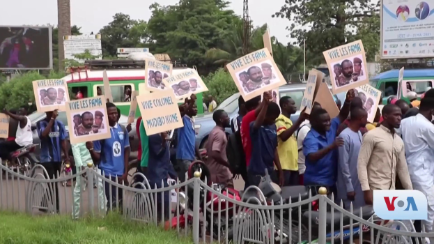Eine Gruppe von Menschen hält Schilder in Händen vor einem Zaun, umgeben von Fahrzeugen, Bäumen, Masten, Werbetafeln und einem Bildschirm, mit Gras unten und dem Himmel im Hintergrund, scheinbar protestieren sie gegen eine Regierungsentscheidung.