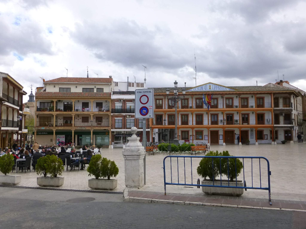 Ein belebter Stadtplatz mit Stühlen und Menschen, umgeben von Topfpflanzen, Metallabsperrungen, Straßenlaternen mit Flaggen, einem Schild, Gebäuden mit Fenstern und einem bewölkten Himmel.