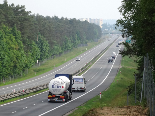 Ein großer Lkw fährt auf einer von beiden Seiten mit Bäumen gesäumten Autobahn, mit einem Zaun und Gras auf der rechten Seite und Gebäuden in der Ferne unter einem klaren blauen Himmel.