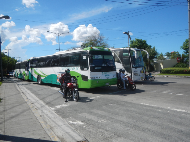 Ein grüner und weißer Shuttlebus steht am Straßenrand mit ein paar Motorrädern davor, ein Fußweg mit Gras und Pflanzen links daneben und Gebäude, Bäume, Laternenmasten und einen klaren blauen Himmel im Hintergrund.